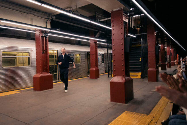 Een man met een donkere jas en donkere broek rent over een ondergronds metroplatform. De man rent van links naar rechts, weg van een zilverkleurige metrotrein die langs het platform staat. Het platform wordt verlicht door TL-buizen en heeft rode stalen pilaren. Rechts van de man staan mensen aan de rand van het platform, en enkele handen klappen in de rechterbenedenhoek van het beeld.