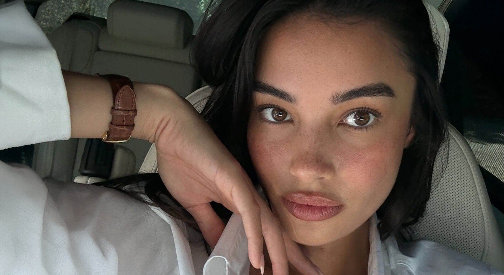Close-up portret van een vrouw met lang donker haar die in een auto zit. Ze draagt een wit overhemd en een horloge met een bruine leren band. Ze kijkt direct naar de camera met een natuurlijke make-uplook met zachtroze blush en lipkleur, waardoor haar sproeten zichtbaar zijn. Ze leunt met haar hand, met de vingers voor haar kin, tegen haar gezicht.