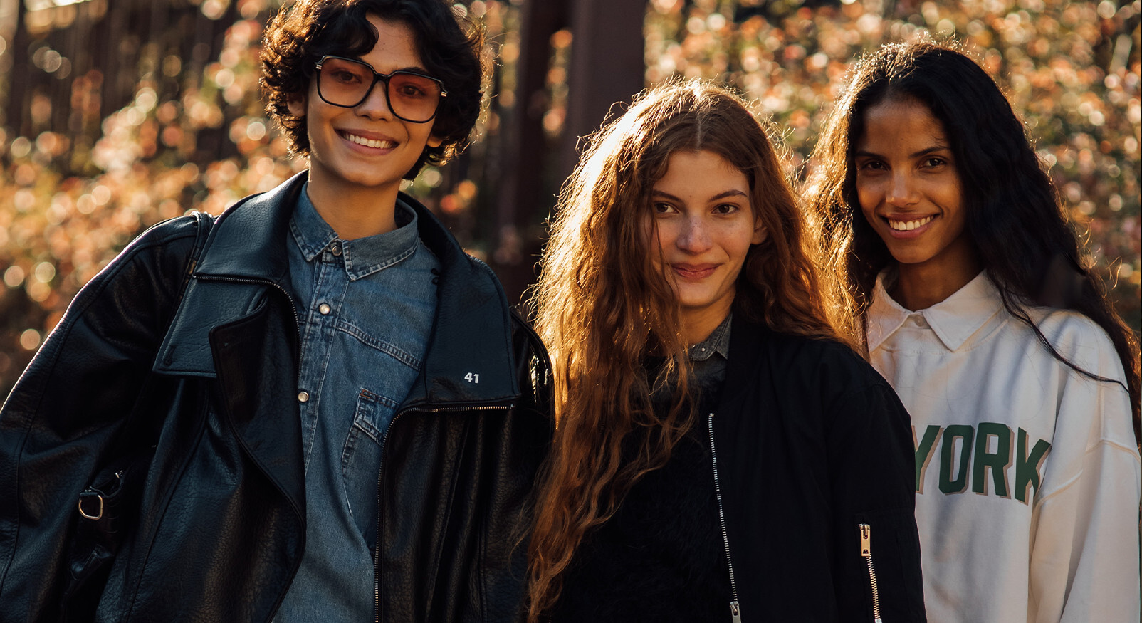 "Drie lachende jonge vrouwen in casual mode, waaronder een leren jas en een New York sweatshirt, poseren buiten in het warme zonlicht."