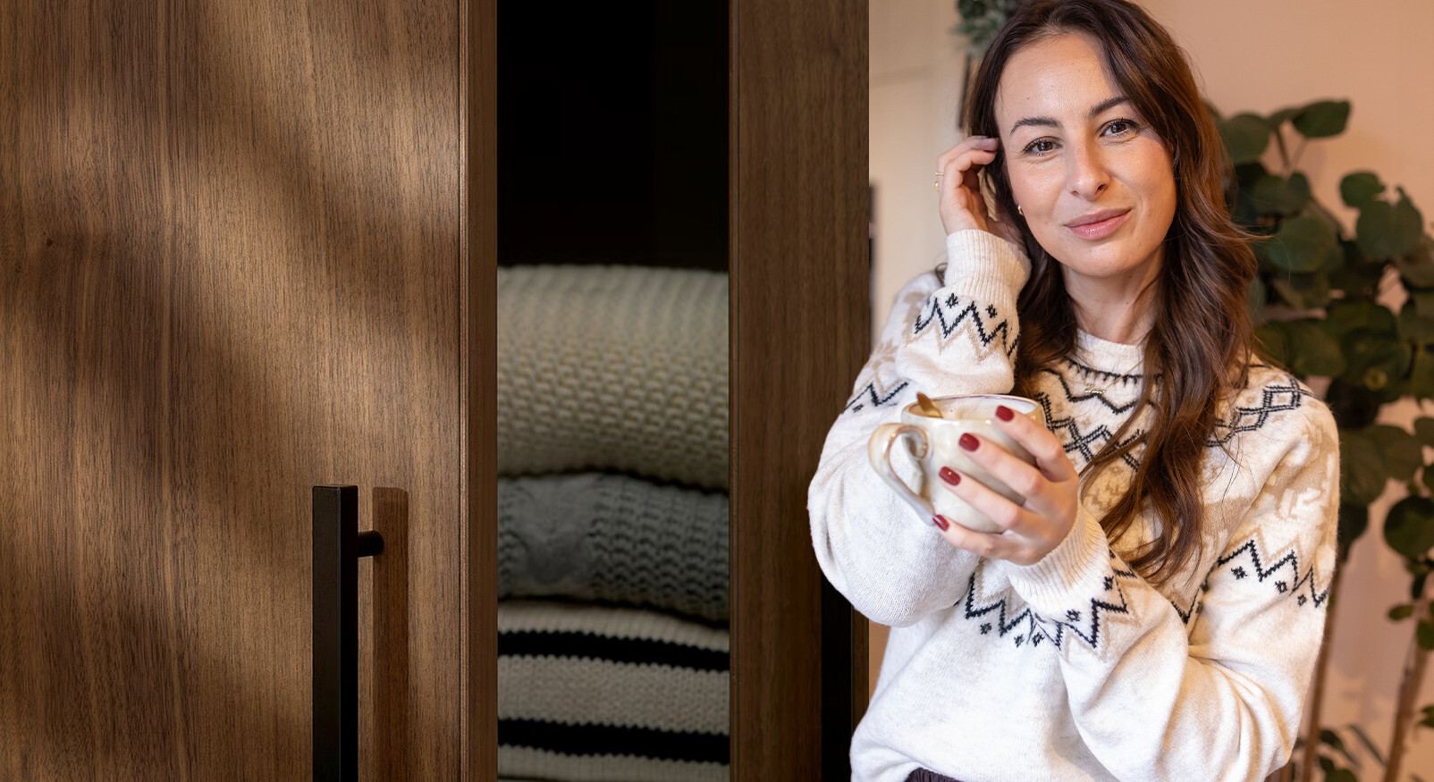 "Smiling woman in a warm winter sweater holding a mug next to a wooden cabinet with blankets."