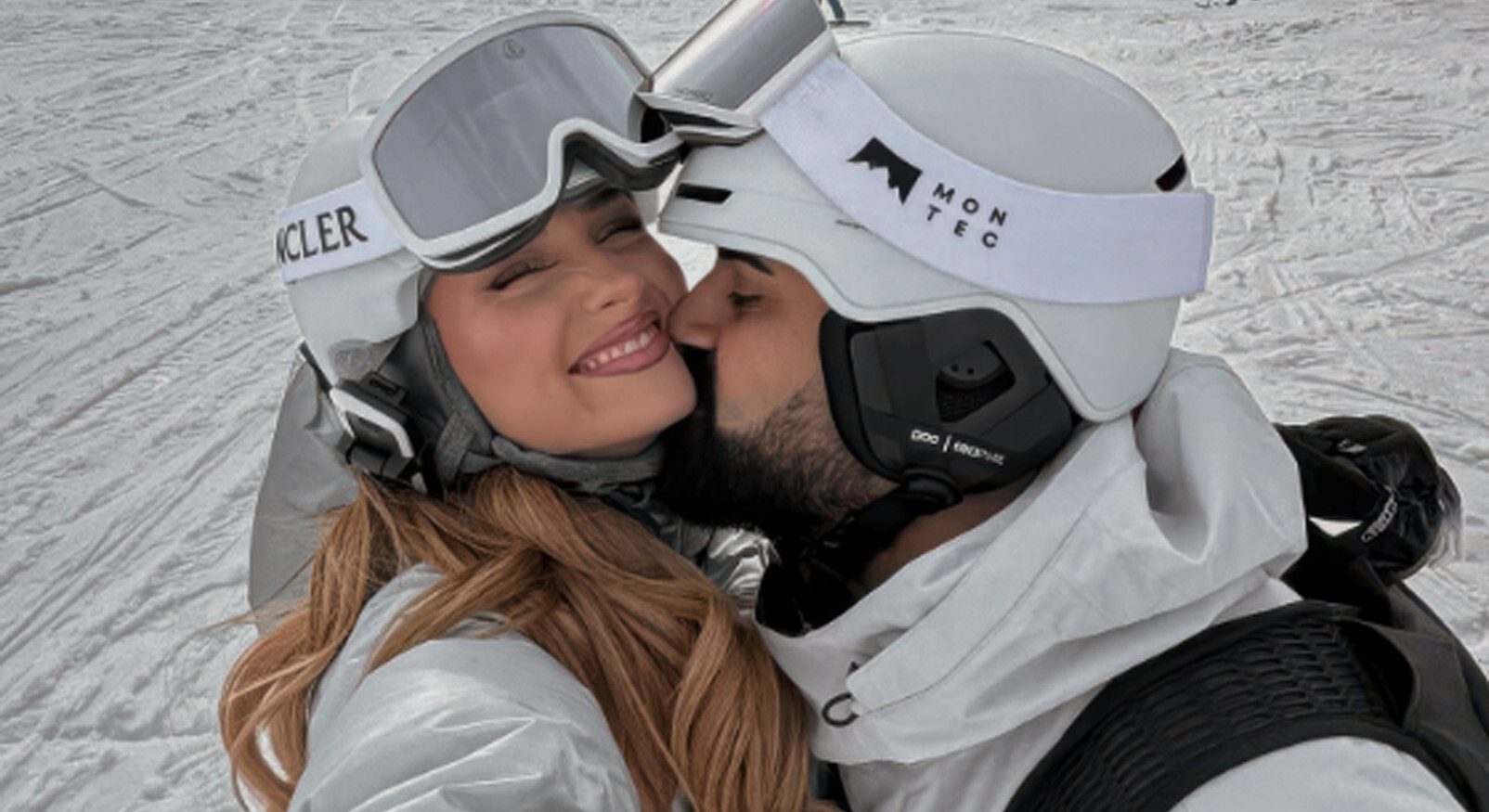 Lachend koppel in witte wintersportkleding met skihelmen en skibrillen op een besneeuwde piste