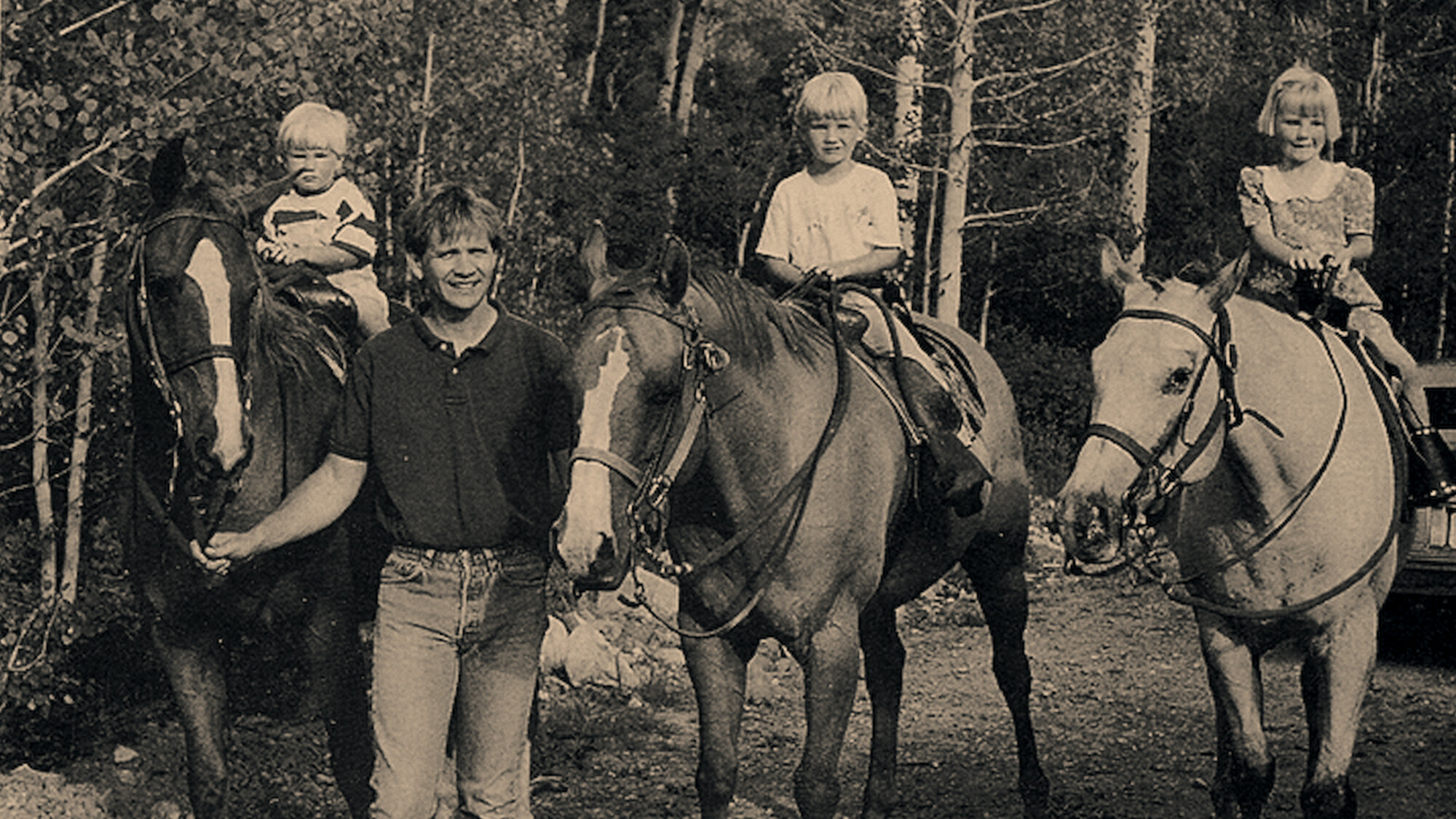 "Zwart-wit vintage foto van een man die drie jonge kinderen op paarden begeleidt in een bosrijke omgeving."