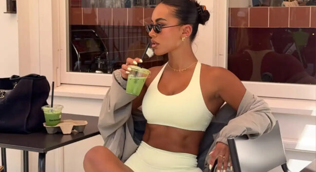 Een jonge vrouw in een lichtgele sportoutfit zit op een terras en drinkt een groen sapje. Ze draagt een zonnebril en heeft een grijze jas over haar schouders. De sfeer is sportief en modieus.