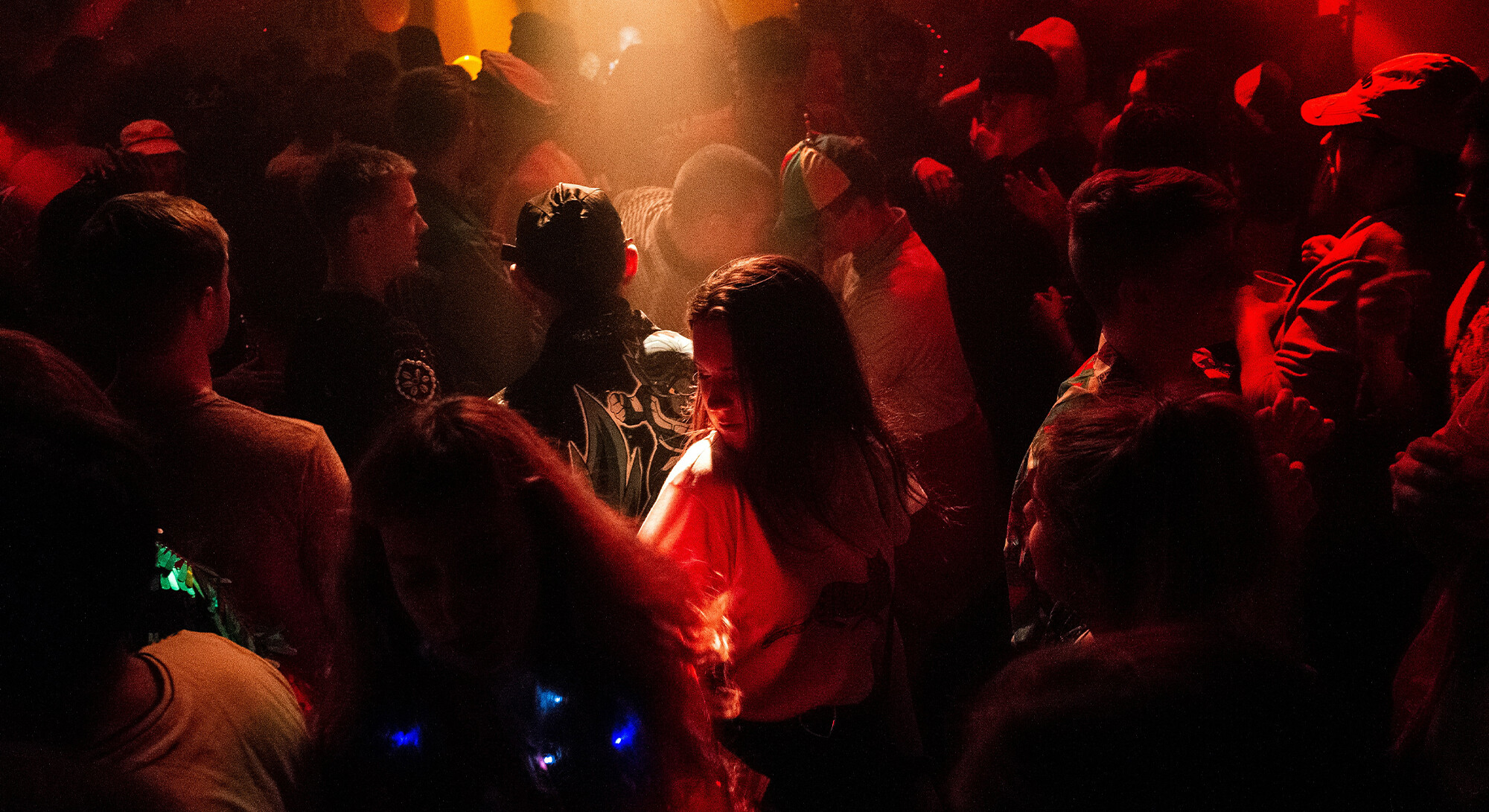 Een sfeervolle, donkere opname van een drukbezochte club of feestlocatie. De ruimte is gevuld met dansende mensen en wordt verlicht door warm, roodachtig sfeerlicht en enkele felle lichtspots. De menigte bestaat uit diverse personen, waarvan sommigen petten of kleurrijke accessoires dragen, wat zorgt voor een dynamische en intieme uitgaansvibe.