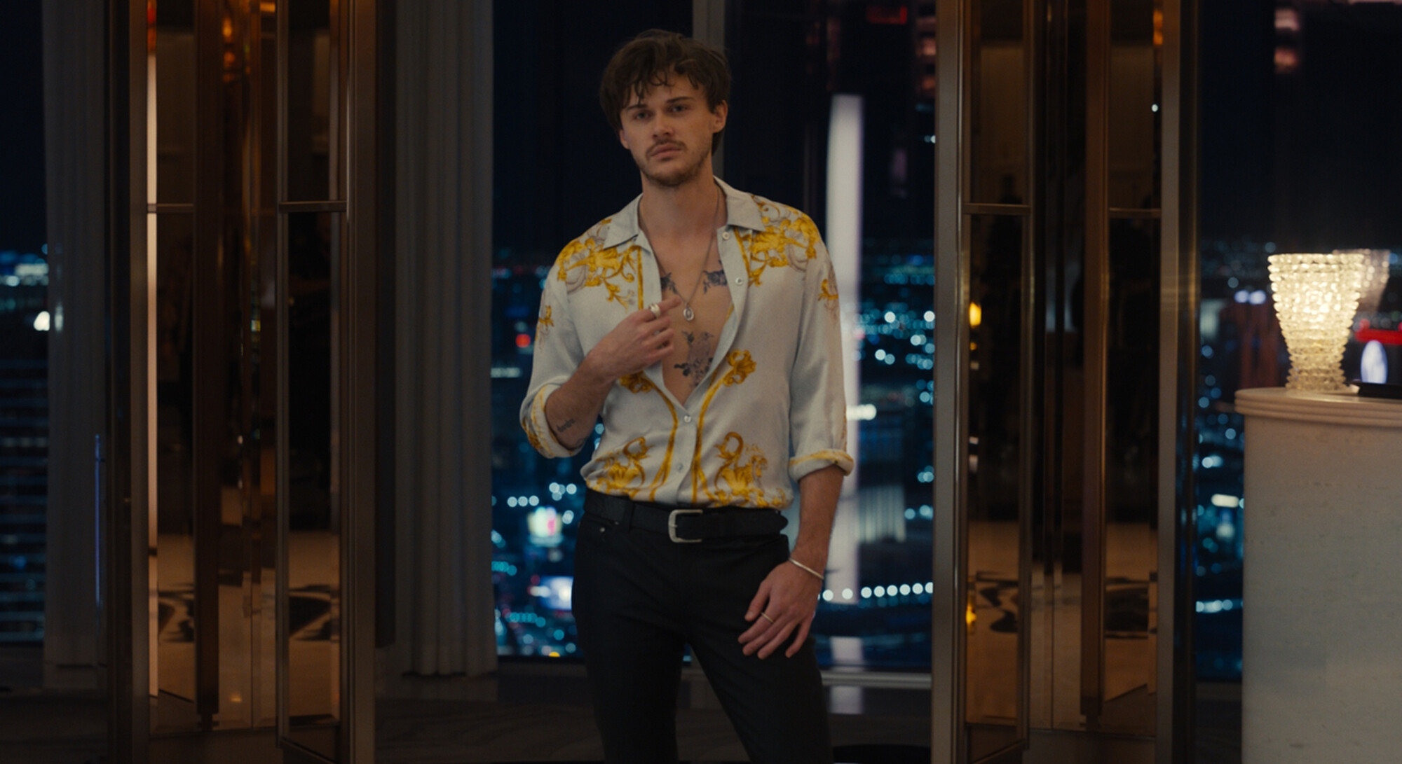 Een jonge man met donker, warrig haar staat in een deuropening van een luxe penthouse met uitzicht op een stad bij nacht. Hij draagt een lichtblauwe blouse met goudkleurige barokke prints die half opengeknoopt is, waardoor tatoeages op zijn borst zichtbaar zijn.
