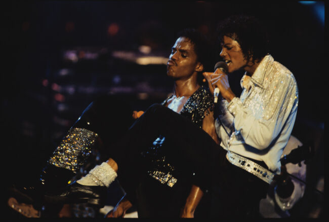 Michael Jackson sings during a performance next to a dancer, dressed in a sparkling white blouse with black pants and glitter boots. 