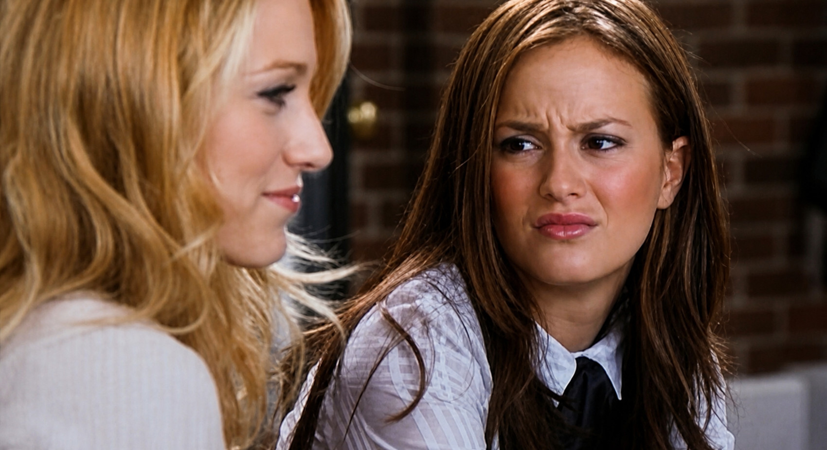 Two young women sit next to each other, with one looking at the other with a skeptical gaze.