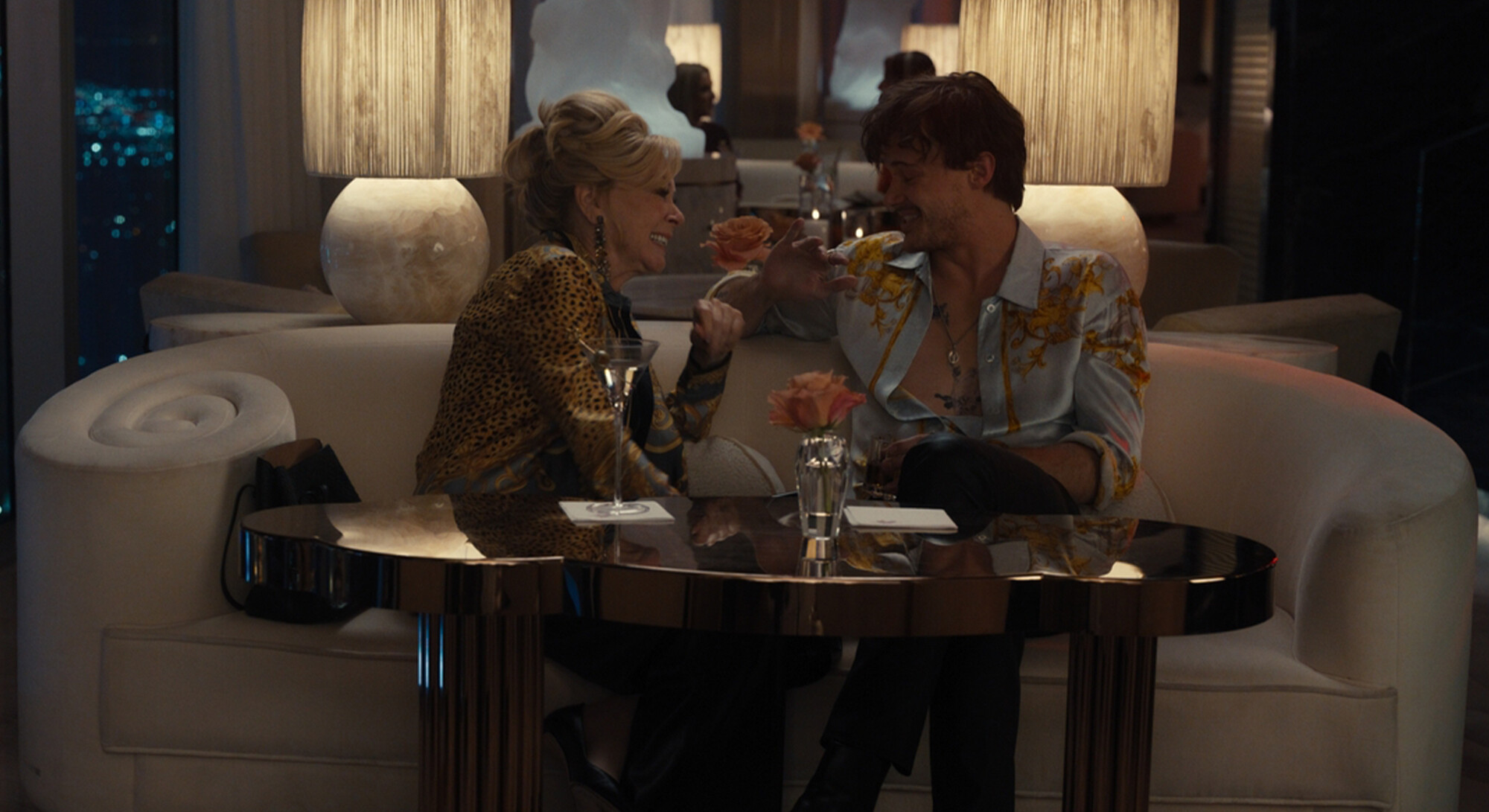 Een jonge man en een oudere vrouw zitten lachend naast elkaar op een witte ronde bank in een chique lounge. De vrouw draagt een jasje met luipaardprint en de man draagt dezelfde blauw-gouden blouse als in de eerste foto. Op tafel voor hen staan drankjes en bloemen.