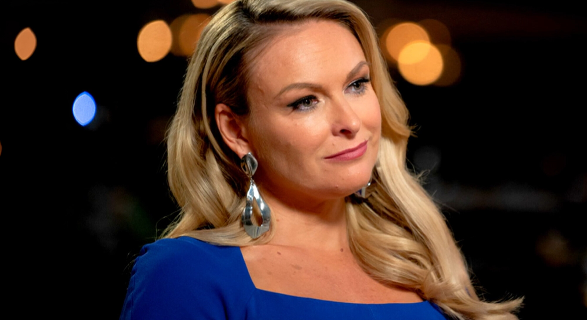 Mel Schilling, bekend als expert van Married at First Sight, kijkt glimlachend opzij tijdens een interview. Ze draagt een blauwe top en opvallende zilveren oorbellen tegen een donkere achtergrond.