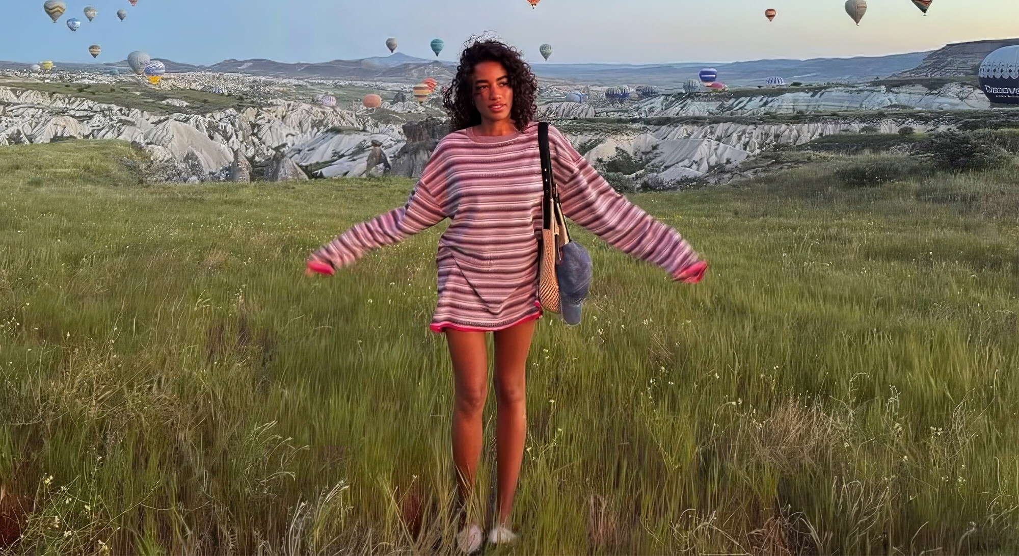 A woman stands with her arms outstretched in a green field in Cappadocia, with rock formations and dozens of hot air balloons in the background.