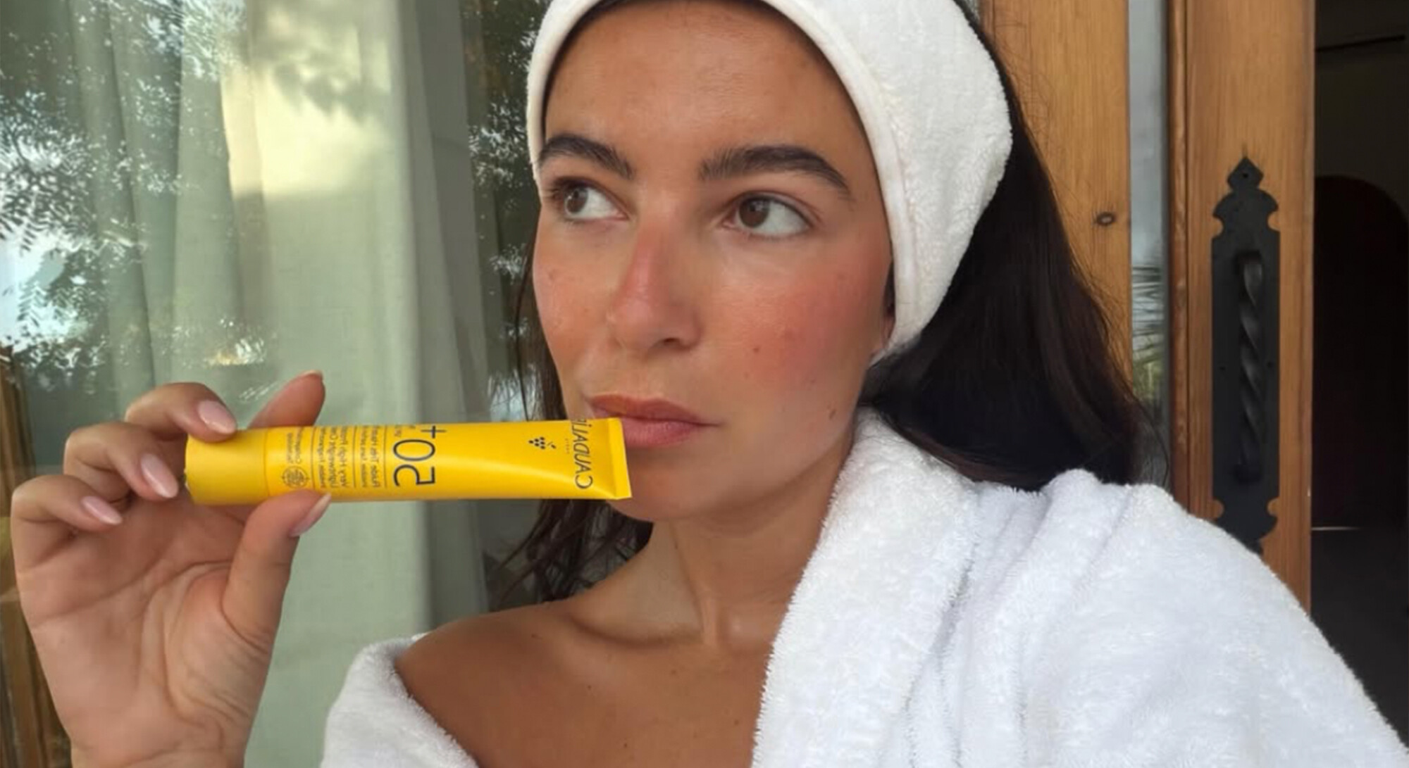 Vrouw in een witte badjas en haarband houdt een gele SPF 20-zonnecrème voor haar gezicht.
