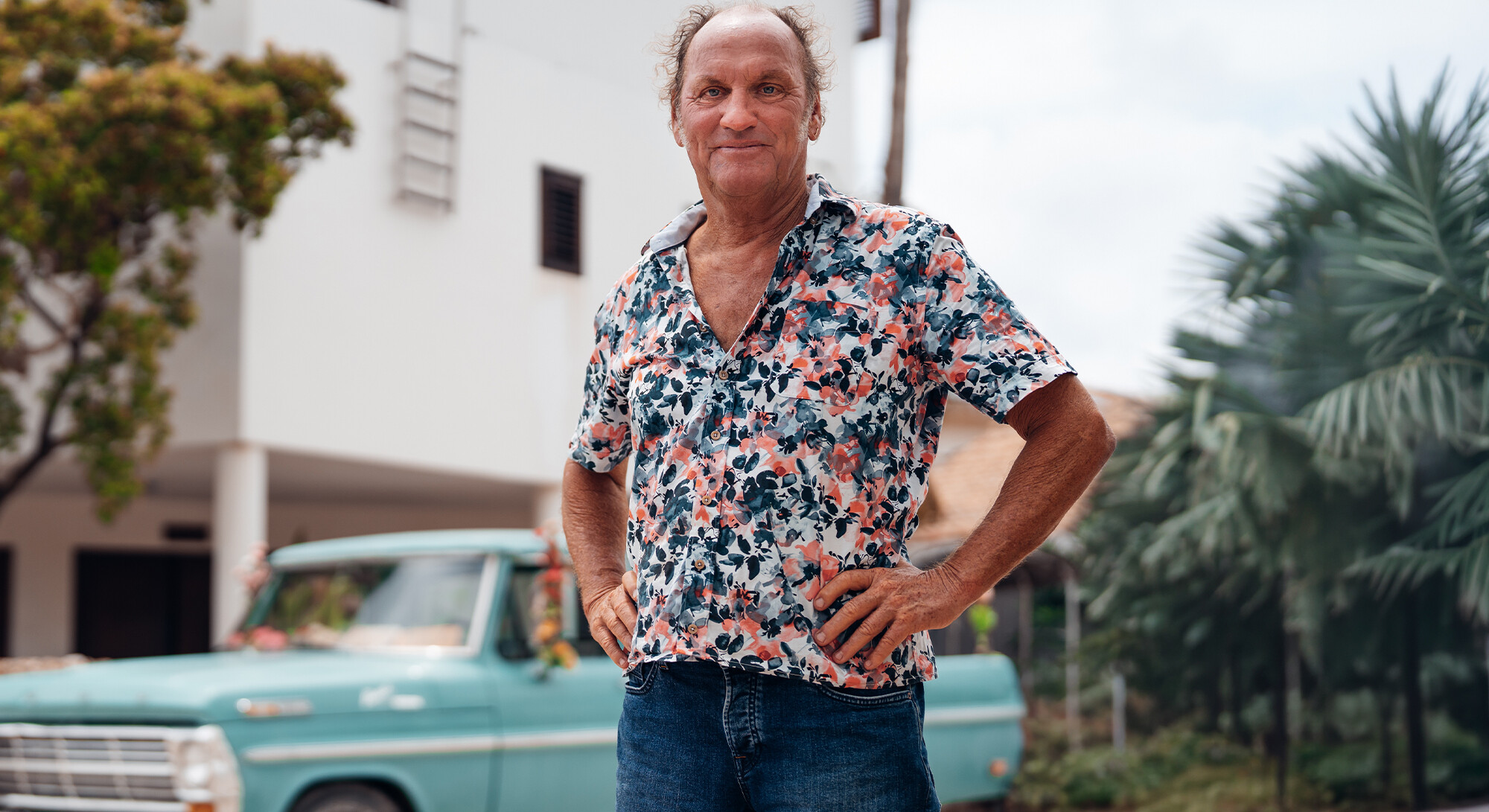 Fred poseert in een gebloemd overhemd voor een witte villa, met bomen en een turquoise oldtimer op de achtergrond.