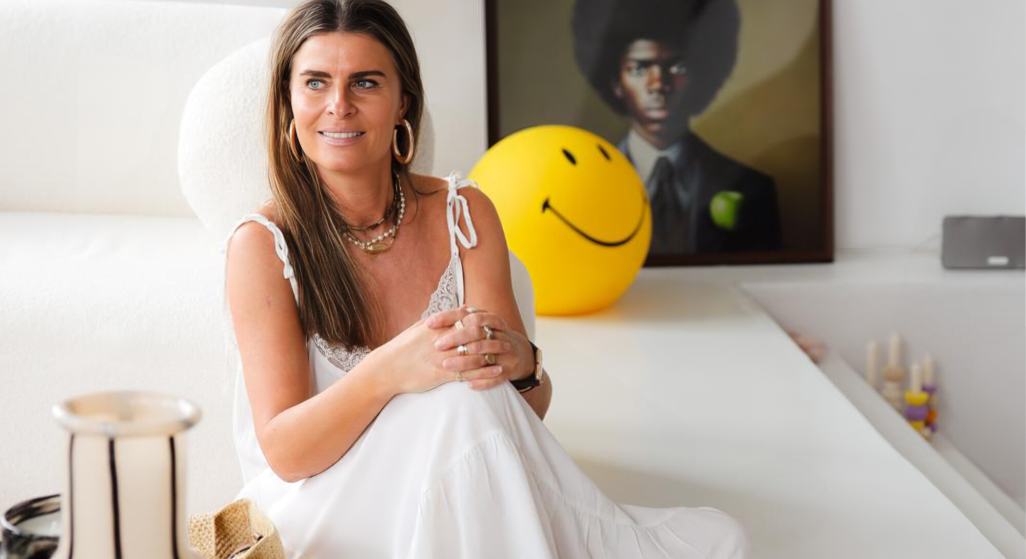 Sabine Noyen is sitting in a white dress in a light living room, with a yellow smiley ball and artwork in the background.