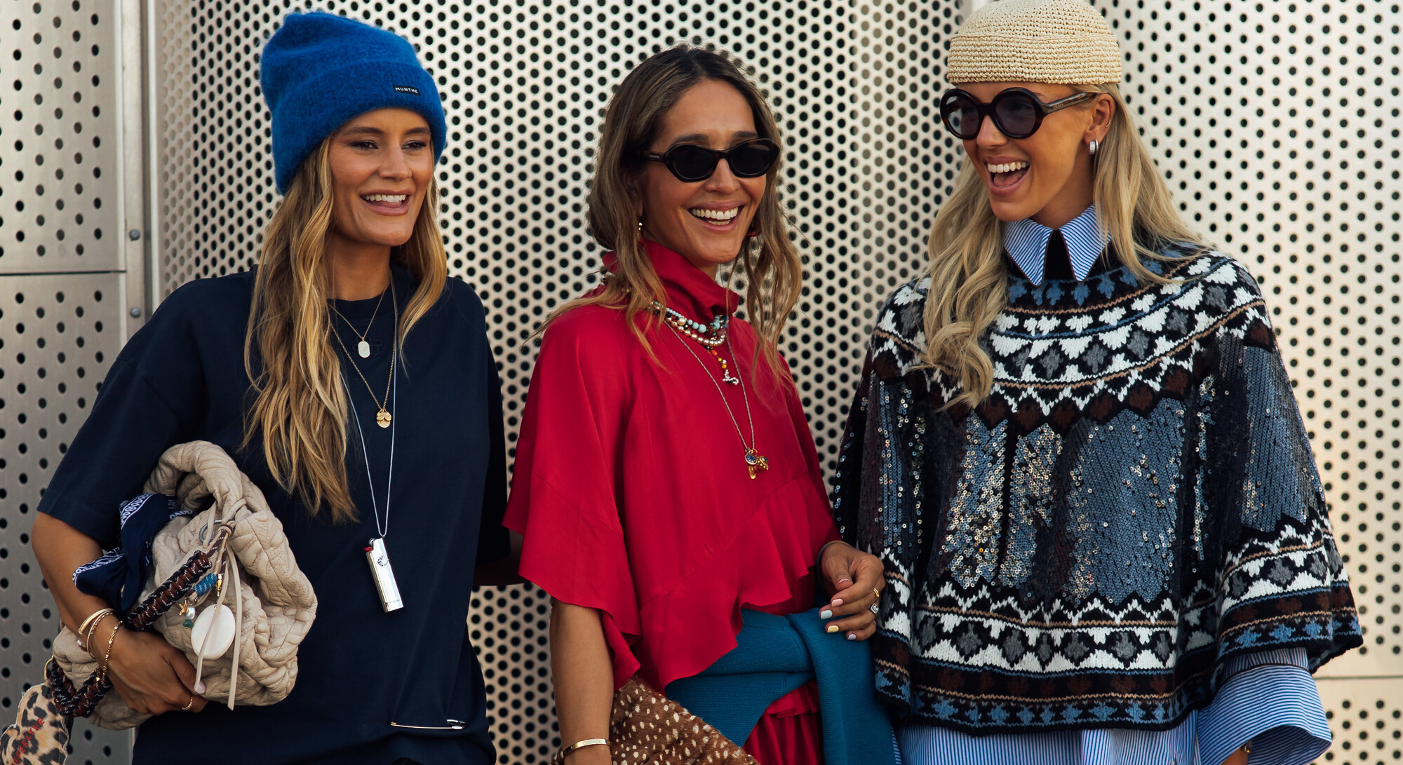 Three smiling women in stylish outfits with hats and sunglasses pose in front of a metal wall.