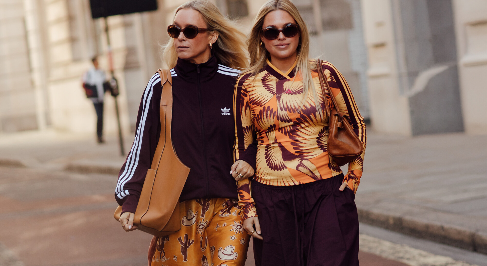 Twee vrouwen met zonnebrillen lopen samen over straat in kleurrijke outfits, waaronder een Adidas trainingsjack en een oranje top met print.