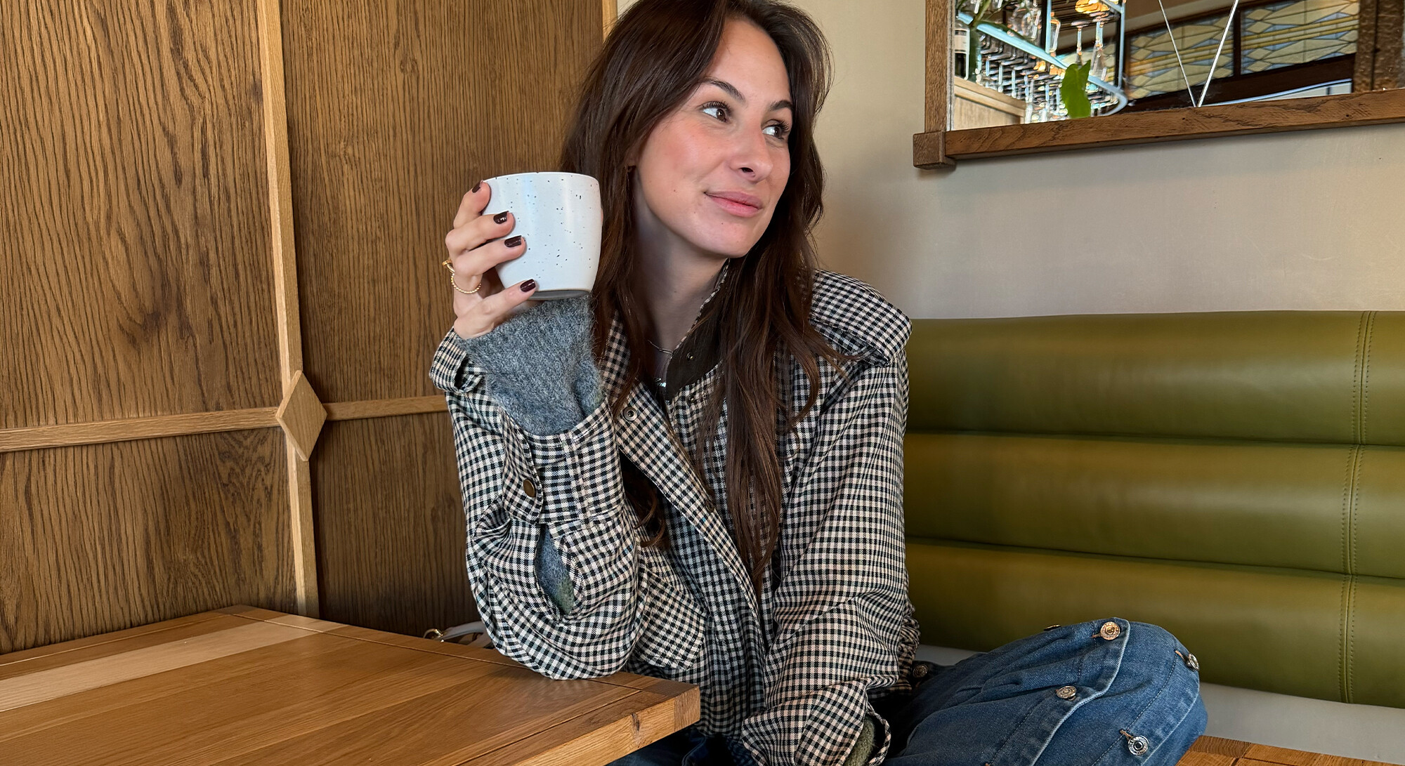 Vrouw in een geruite jas zit aan een houten tafel in een café en houdt een witte mok vast.