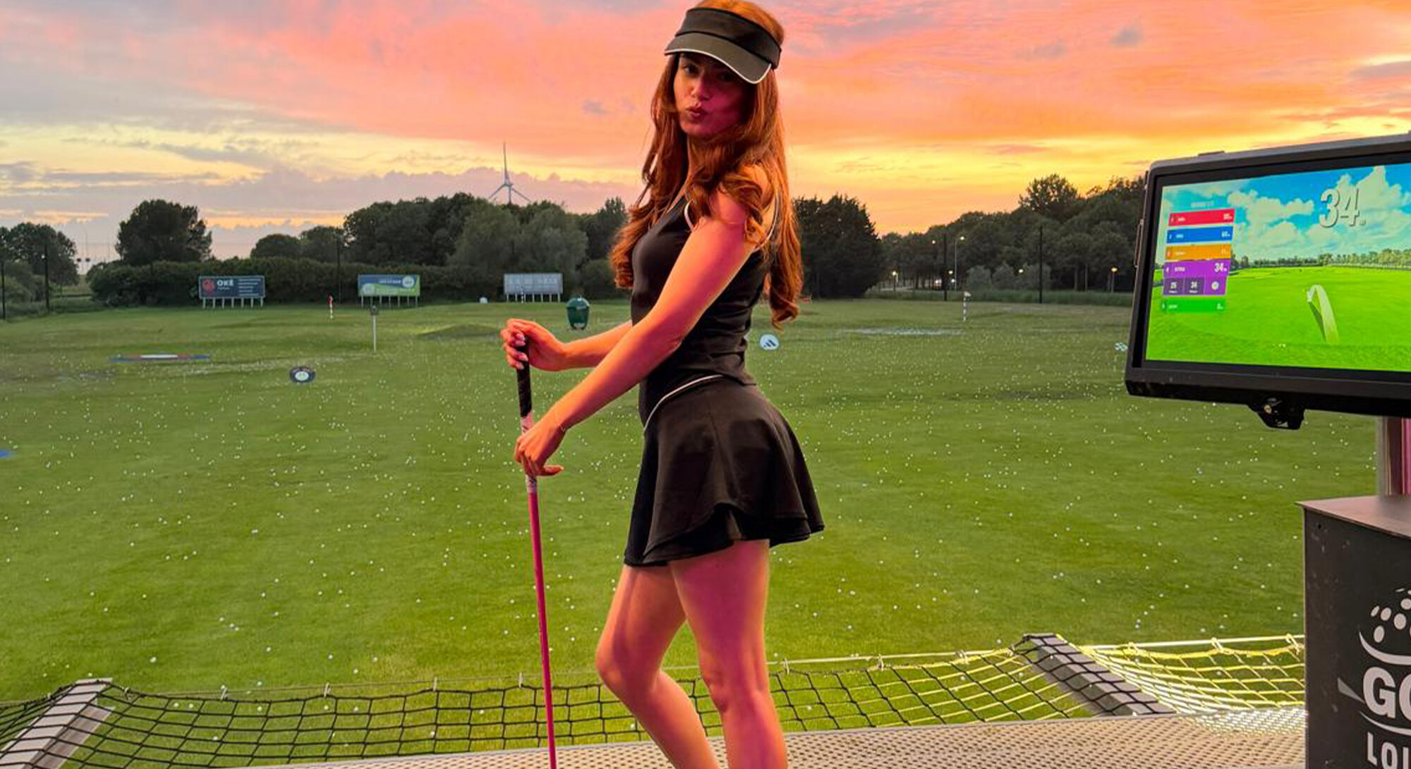 Woman in a black golf outfit poses with a golf club on a driving range during a colorful sunset.