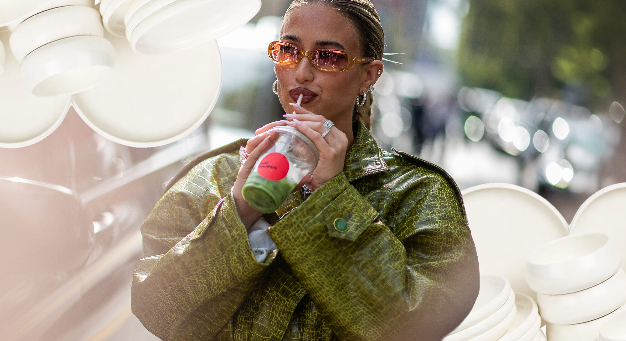 Vrouw in groene leren jas met oranje zonnebril drinkt een groene matcha uit een beker.