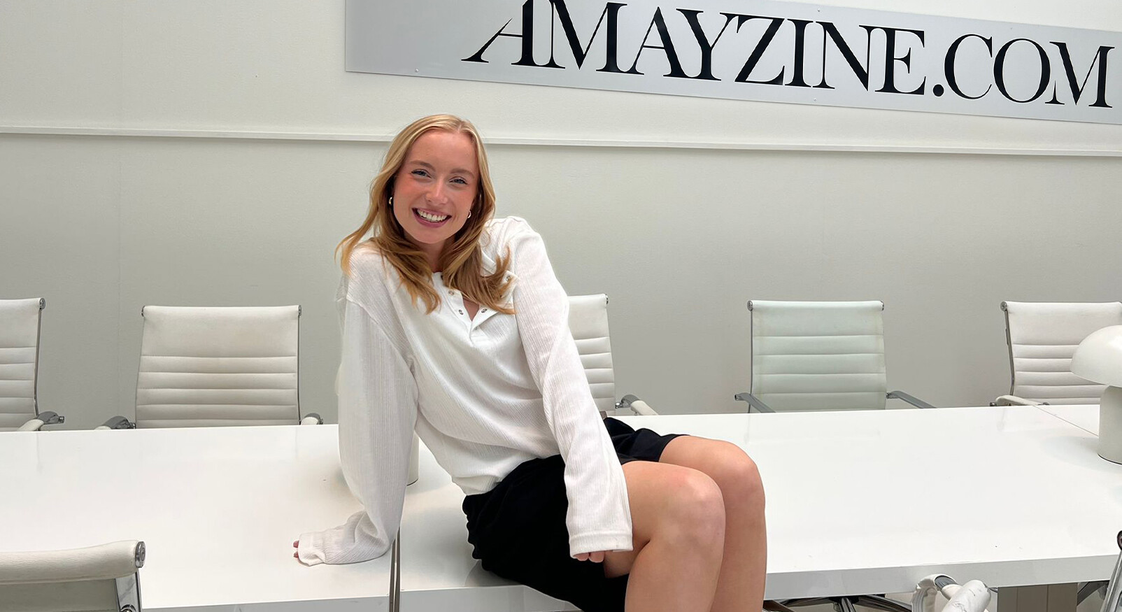 Marit is laughing at a white conference table in the AMAYZINE office, with the AMAYZINE.com logo in the background.