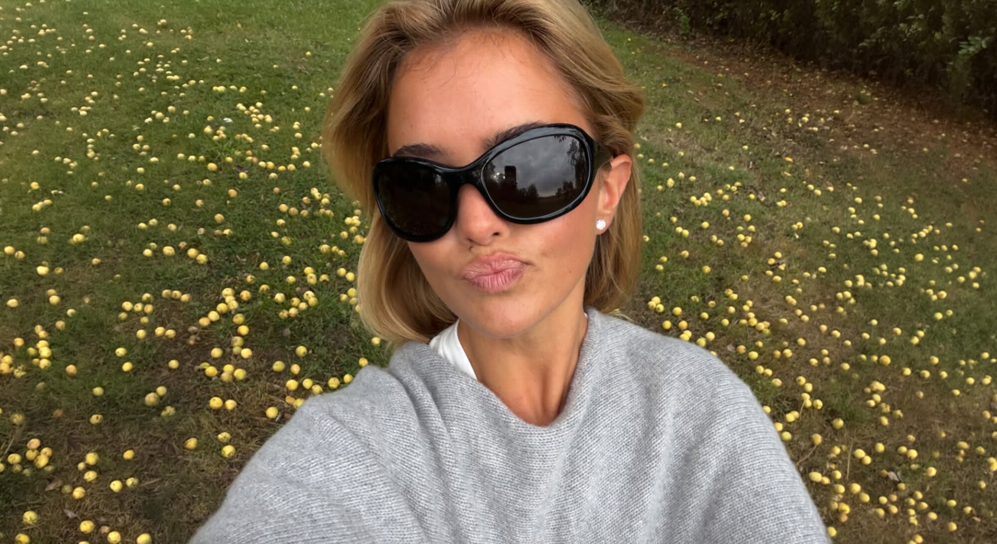 Vrouw met grote zwarte zonnebril en grijze trui maakt een selfie in een boomgaard met op de grond tientallen gele appels.