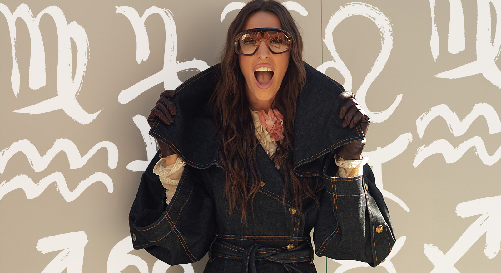 A woman in a stylish outfit poses energetically against a backdrop featuring astrological symbols. She is wearing oversized sunglasses, a dark denim coat with a large collar, brown gloves, and a floral scarf. Her long hair flows down, and she appears to be laughing or shouting joyfully. The overall theme is fashionable and vibrant.