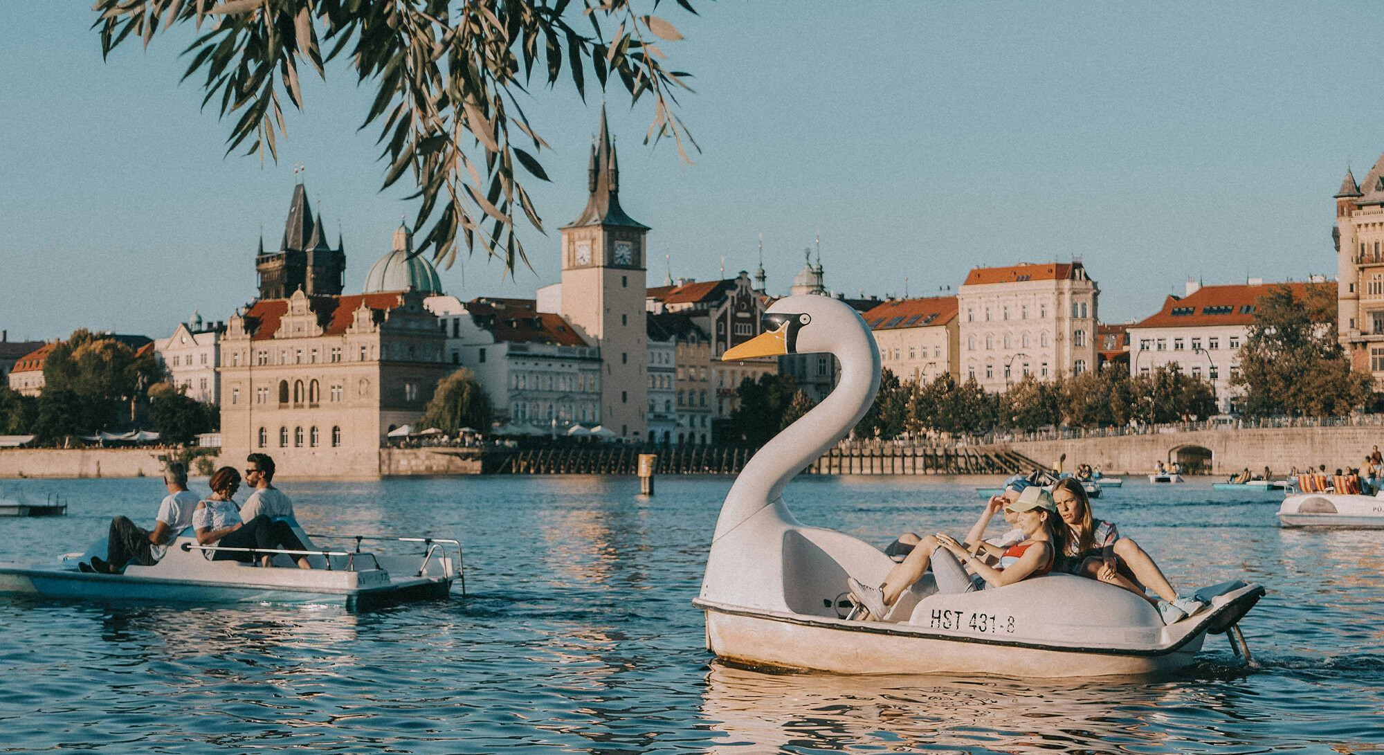 Zwaanvormige waterfiets met passagiers op een rivier, met andere waterfietsen en historische gebouwen met torens op de achtergrond.