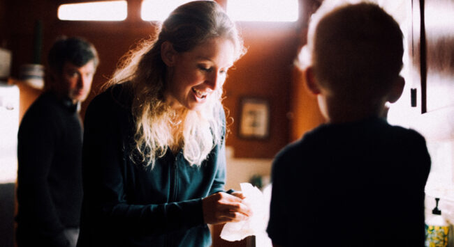 Een vrouw met lang blond haar lacht vrolijk terwijl ze naar beneden kijkt naar een kind dat wazig op de voorgrond staat. Ze houdt iets wits vast in haar handen, terwijl een man op de achtergrond toekijkt in een warm verlichte kamer.