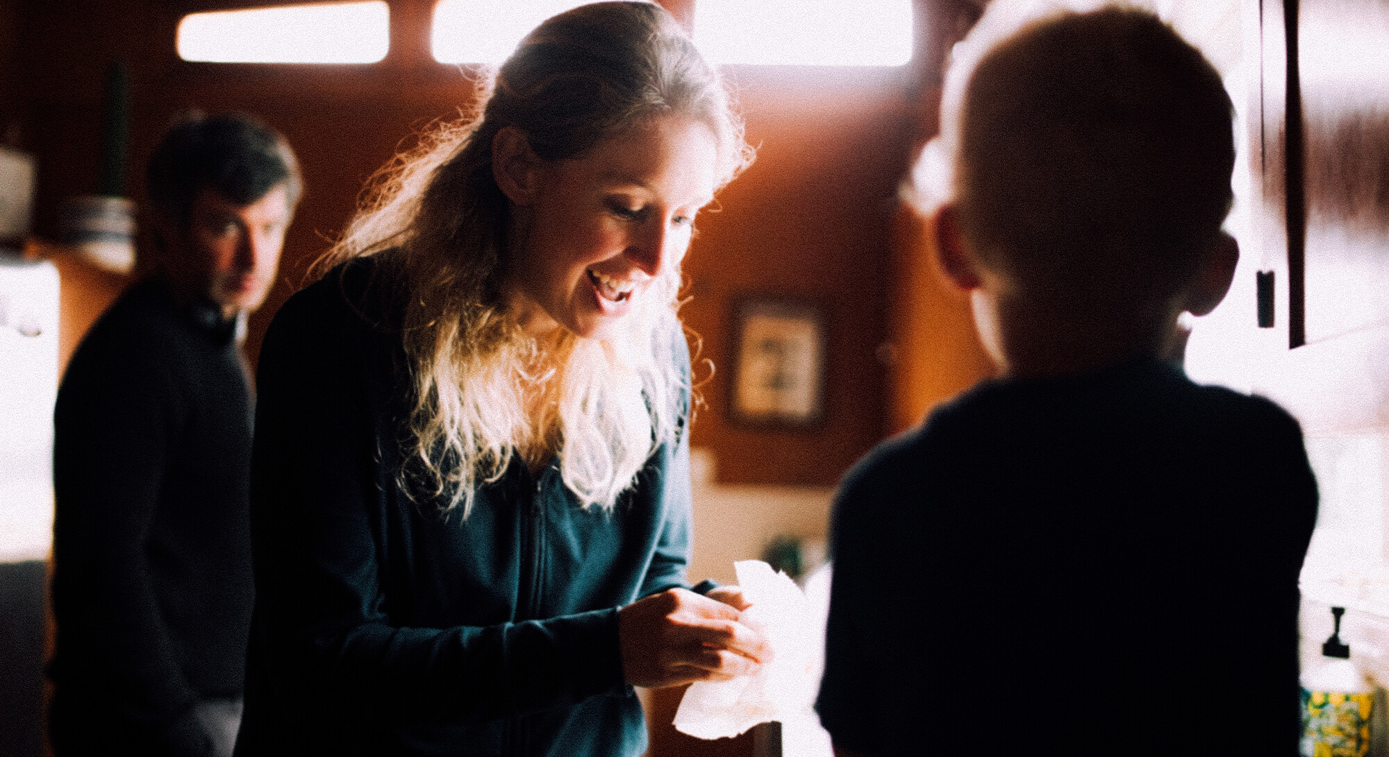 Een vrouw met lang blond haar lacht vrolijk terwijl ze naar beneden kijkt naar een kind dat wazig op de voorgrond staat. Ze houdt iets wits vast in haar handen, terwijl een man op de achtergrond toekijkt in een warm verlichte kamer.