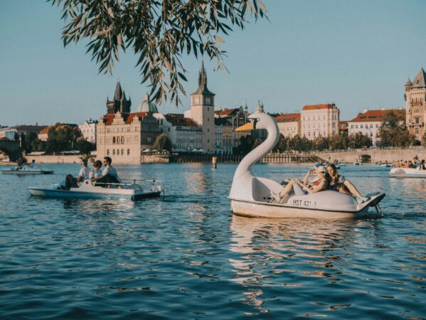 Zwaanvormige waterfiets met passagiers op een rivier, met andere waterfietsen en historische gebouwen met torens op de achtergrond.