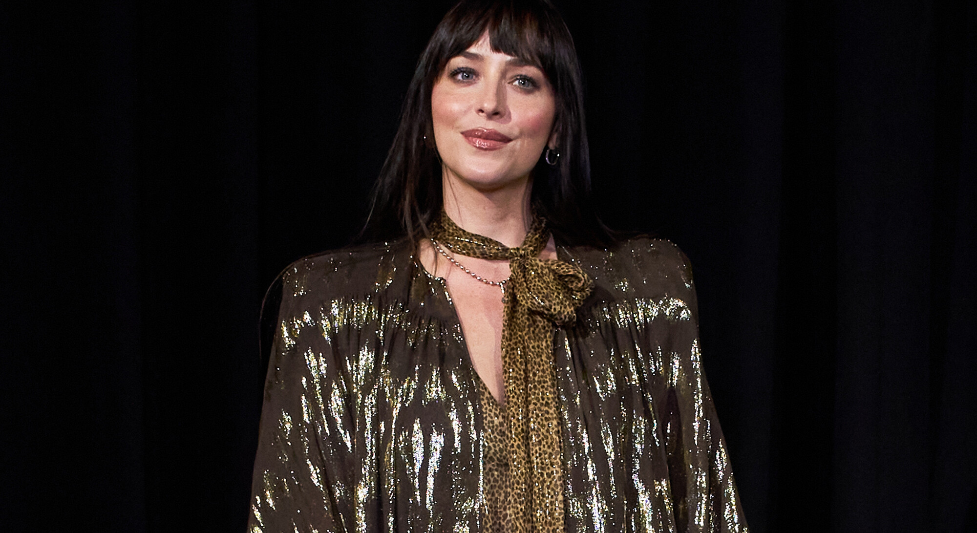 Dakota Johnson met een donkere pony, een glanzende blouse en een sjaal met panterprint tegen een zwarte achtergrond.