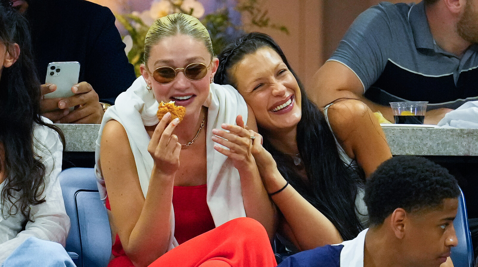 Gigi en Bellla Hadid heel hard lachen en eten tijdens wedstijd kijken zonnebril rode outift